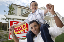 Hispanic Father and Son in Front of Their New Home with Sold Home For Sale Real Estate Sign.